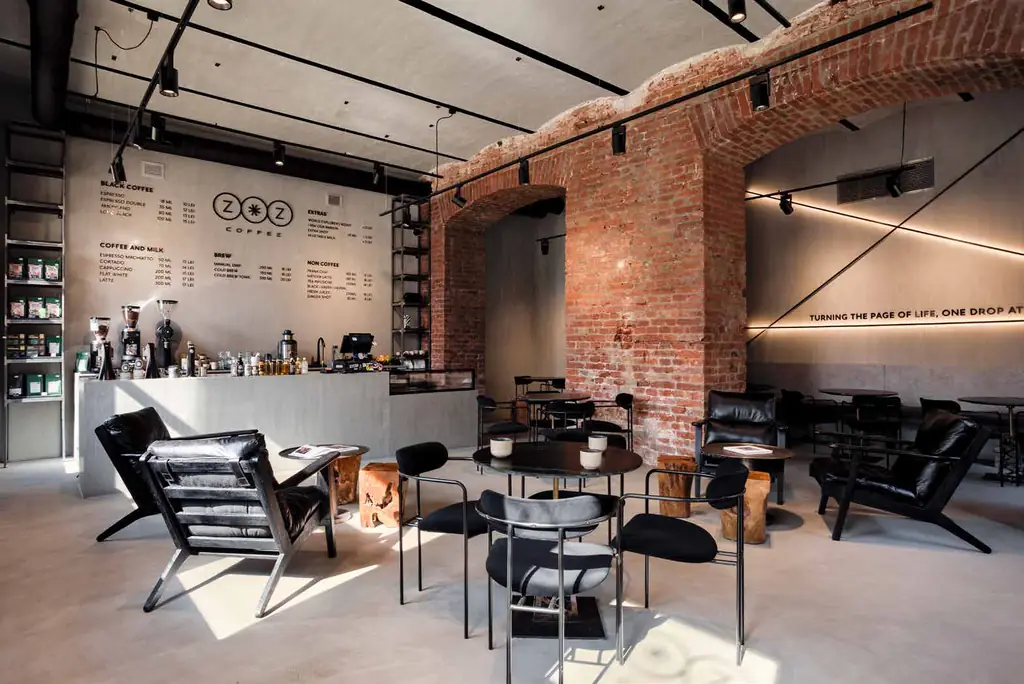 Vedere largă a cafenelei: tejgheaua gri cu meniul pe perete, arcadele din cărămidă și fotolii negre în lumina zilei.